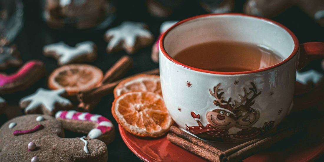 Mug with reindeer design filled with a warm festive tea, surrounded by gingerbread cookies, dried orange slices, and cinnamon sticks on a cozy table.