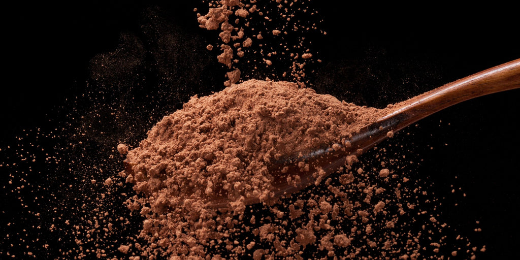 A wooden spoon is scattering organic cocoa powder against a black background, highlighting the dynamic texture and motion.