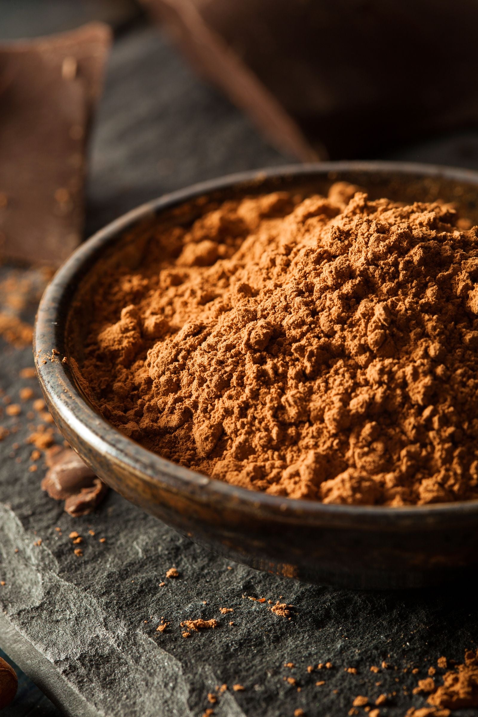 Close-up of organic cacao powder in a rustic bowl, set against a dark slate background with chocolate pieces.